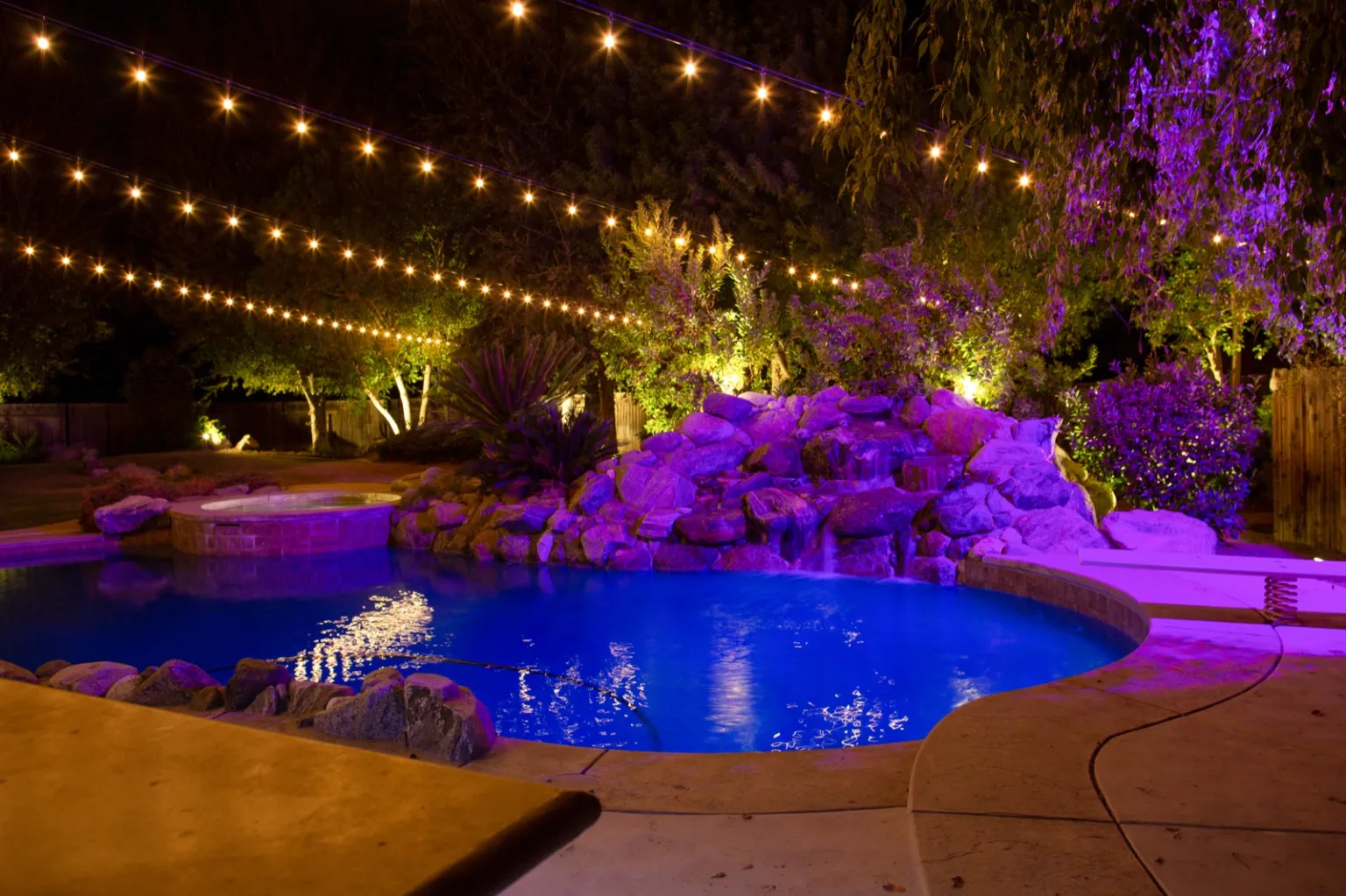 Backyard pool, rock landscaping, trees, and overhead string lights illuminated at night