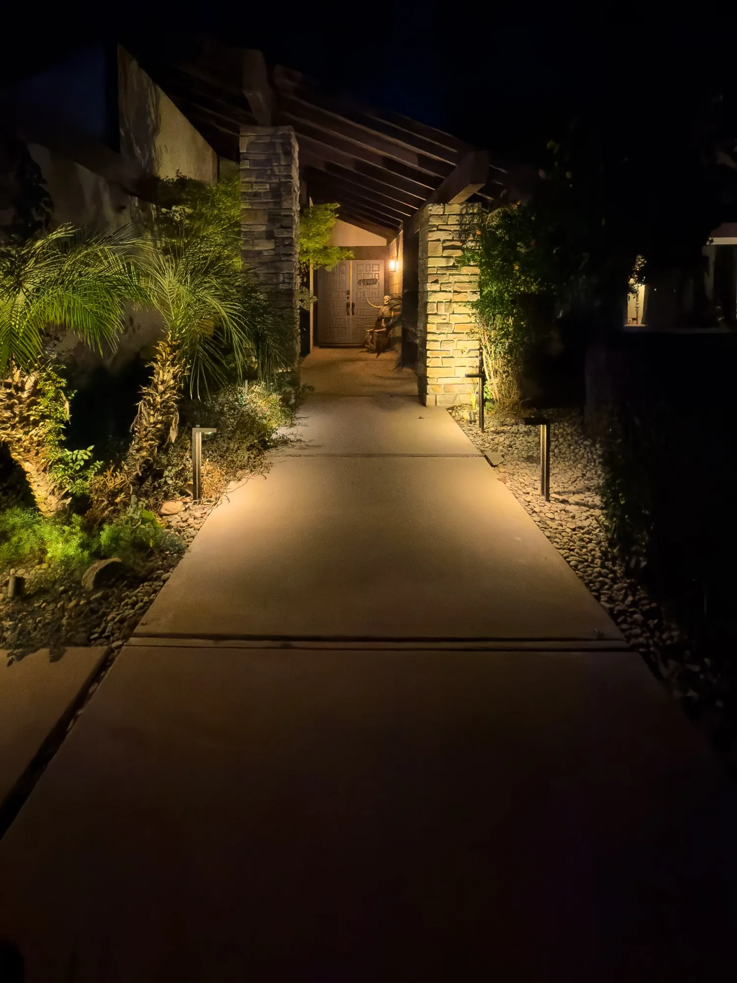 Lit entry walkway with path lights, uplit palms, and stone columns