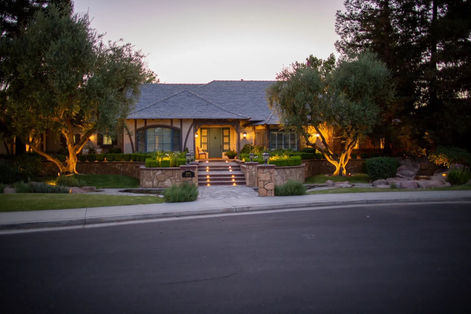 Front elevation at dusk with lit olive trees, stone walls, step lights, and entry lighting